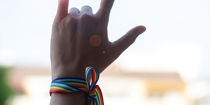 hand in the "I love you" sign in American Sign Language with a rainbow LGBT support ribbon tied around the wrist