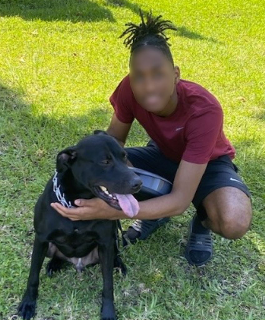 A youth from Brooksville Youth Academy kneeling next to Knox, a black lab. The youth has his hand on the dogs chest and is holding a food bowl. 